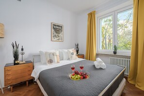 A hotel room with a bed, bedside table, a framed picture on the wall, a window with curtains, and a view of trees outside. - Lumina 2BR Apart near Center of Warsaw (Warszawa)