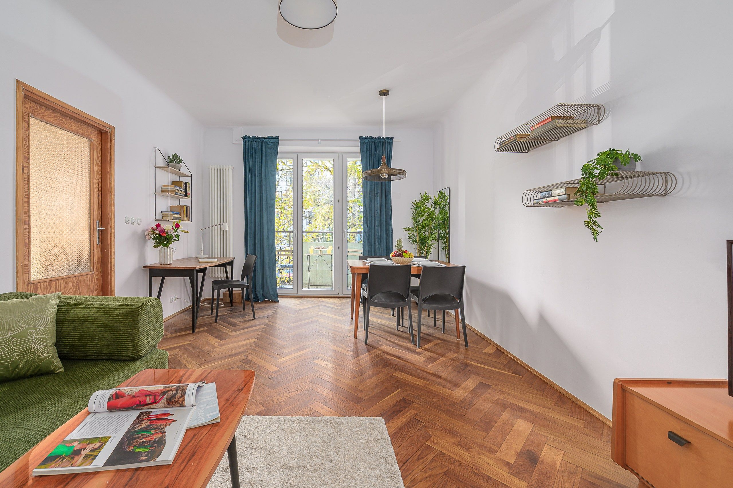A modern living room with a herringbone floor, a green sofa, a dining area with a table and chairs, and a kitchen area with a built-in oven and a hanging plant.