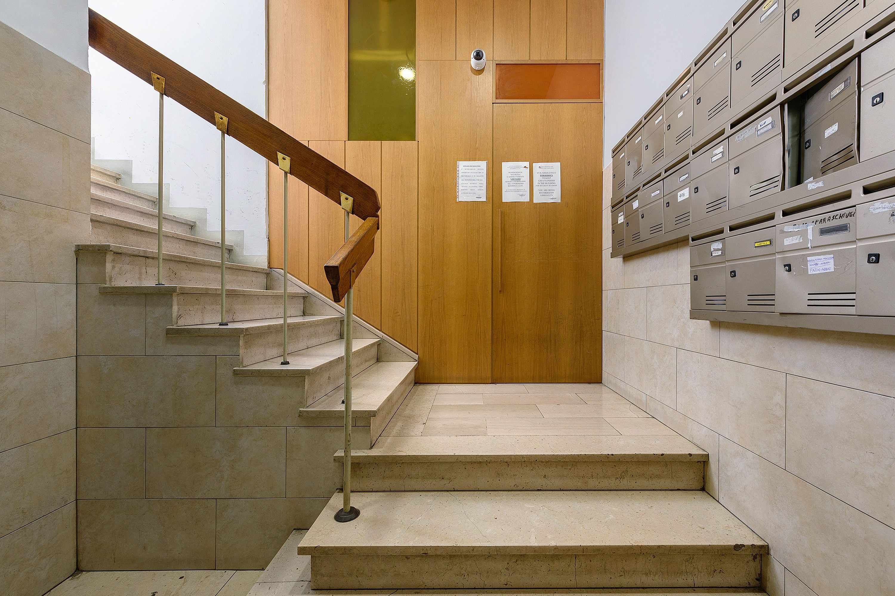 Un escalier mène à une porte en bois sur laquelle est fixée une caméra de surveillance. Plusieurs casiers de rangement sont disposés sur le côté droit.
