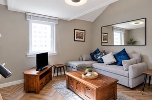 Un salon moderne avec un téléviseur posé sur un meuble en bois, un canapé gris agrémenté de coussins bleus, une table basse en bois et un grand miroir accroché au mur.