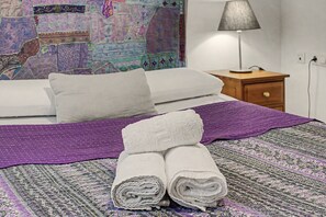 A neatly made bed with a purple blanket and two rolled towels, a wall tapestry, a bedside lamp, and a wooden nightstand. - Apartment 'Galindo II Center Premium Suites' with Private Terrace, Wi-Fi and Air Conditioning (Sevilla)