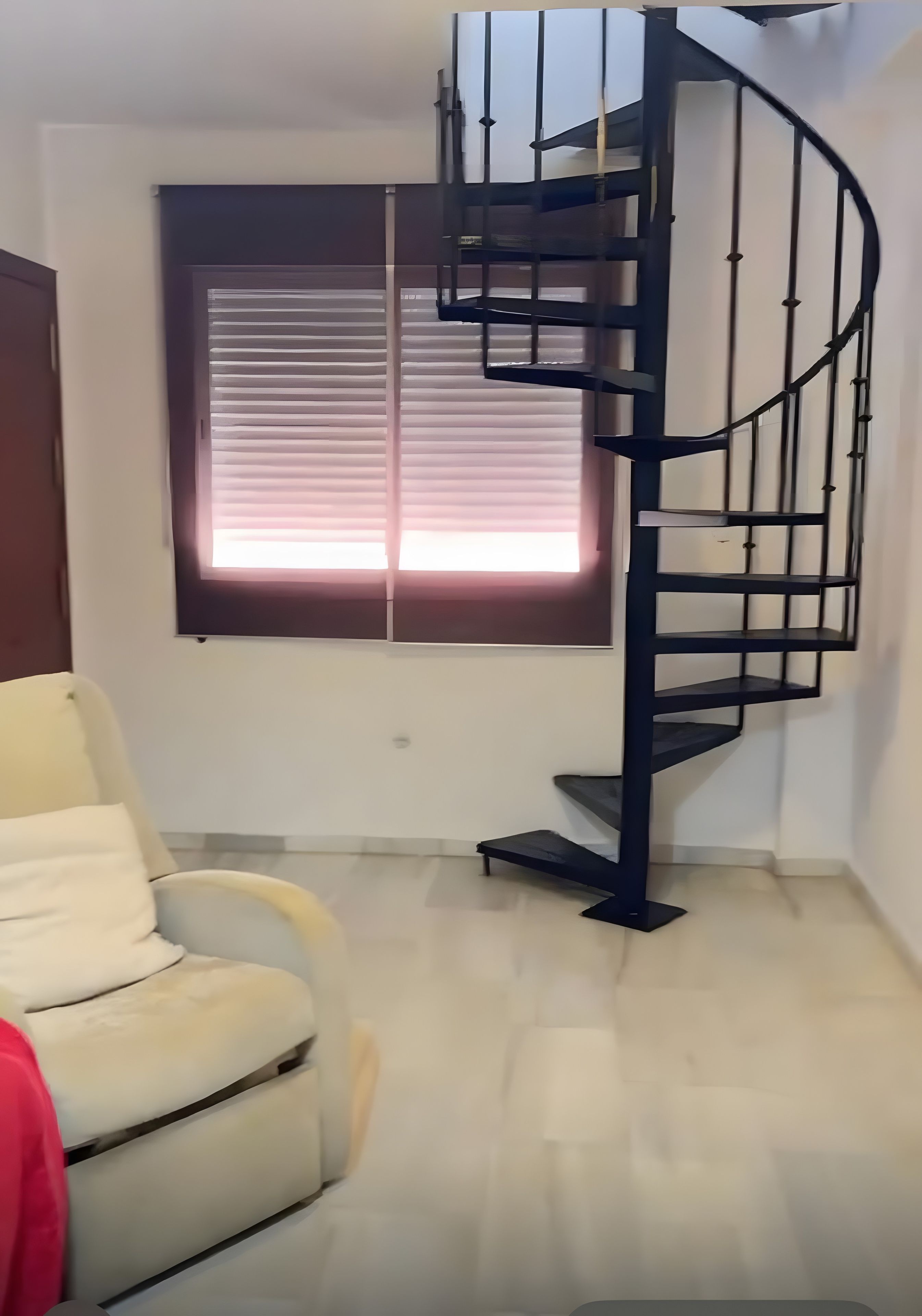A room with a spiral staircase, a window with blinds, and a white armchair.