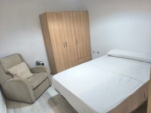 A bedroom with a bed, a wooden wardrobe, a beige armchair, and a small heater. - Apartment 'Macarena Seville Premium Suites' with Private Terrace and Air Conditioning (Sevilla)