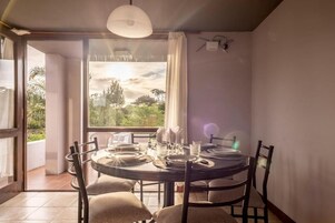 Un comedor con una mesa redonda dispuesta para cuatro personas, un amplio ventanal con vistas a zonas verdes y una lámpara en el techo.