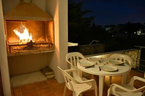 Un área de comedor al aire libre con una parrilla de horno de ladrillo y sillas de plástico blanco.