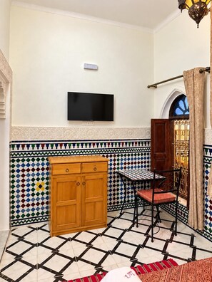 Quarto com piso de azulejos, um armário de madeira, uma cadeira de metal preto e uma televisão fixada na parede.