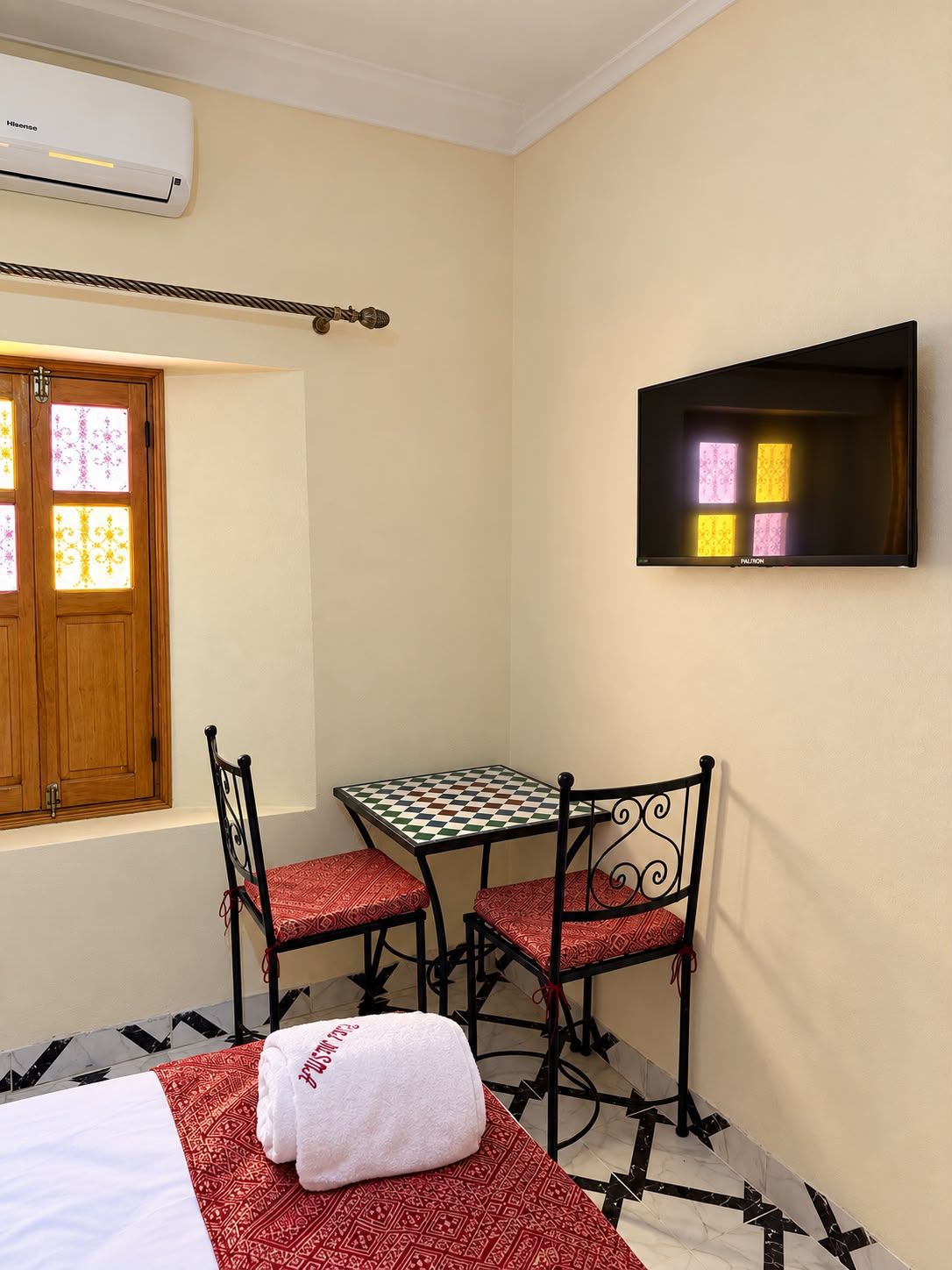 Una habitación pequeña con cama, una mesa y dos sillas, televisión y ventana con vidrios decorativos.