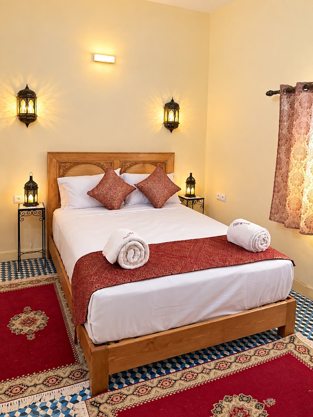Una habitación de hotel con una cama de madera, almohadas estampadas, una colcha rojo y blanco, dos lámparas fijadas a la pared y una alfombra con diseño.