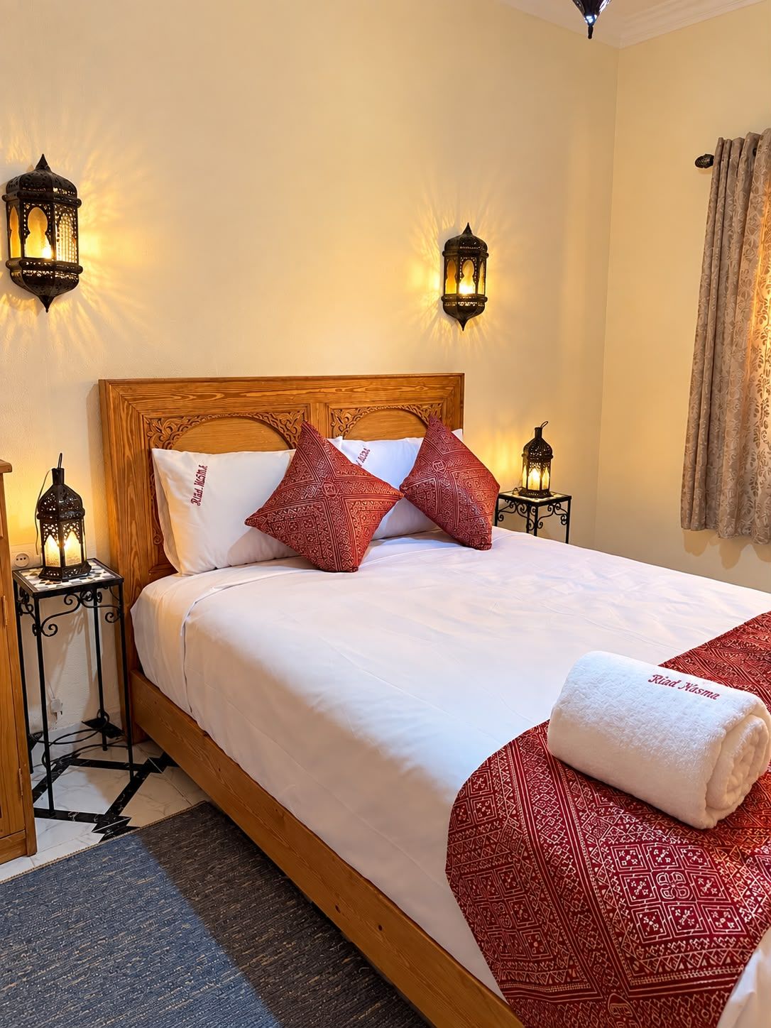 A neatly made bed with a wooden headboard, two red patterned pillows, a white blanket, and a rolled towel with text on it. There are two wall-mounted lanterns on either side of the bed.