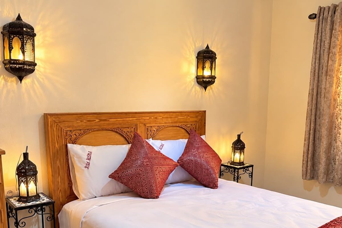 A neatly made bed with a wooden headboard, two red patterned pillows, a white blanket, and a rolled towel with text on it. There are two wall-mounted lanterns on either side of the bed.