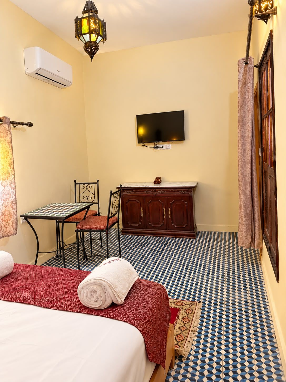 Quarto de hotel com cama, escrivaninha, televisão e piso estampado.