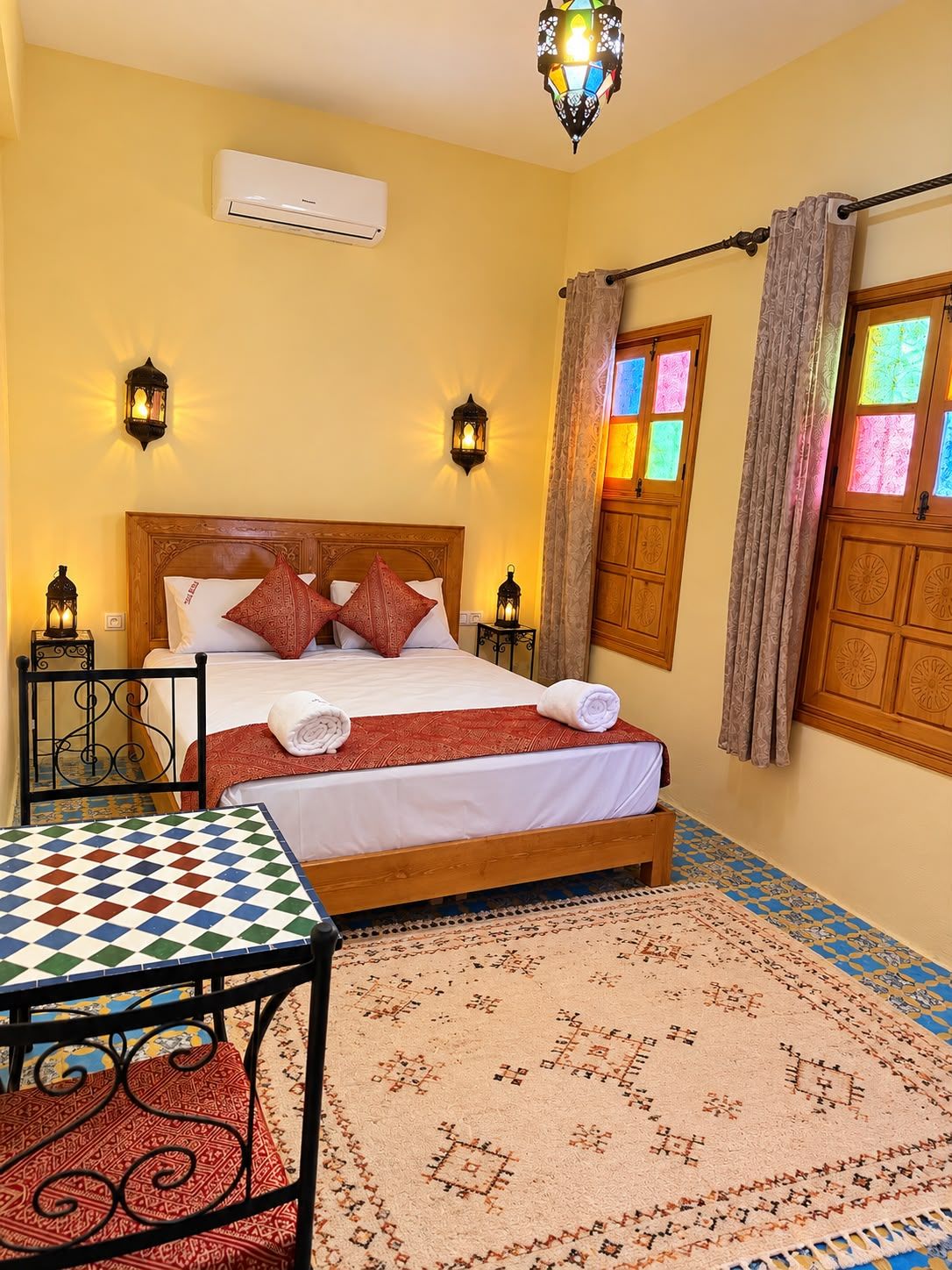 A hotel room with a bed, a table, a patterned rug, and a window with curtains.
