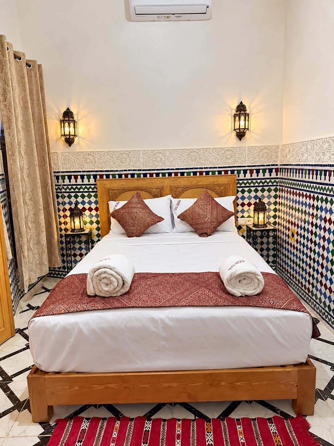 Quarto de hotel com cama, almofadas decorativas, ar condicionado fixado na parede e ladrilhos estampados.