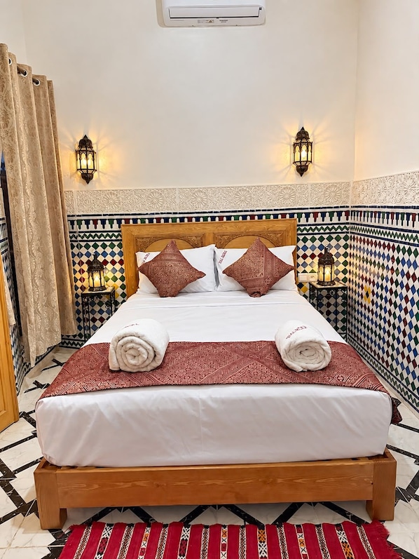 A hotel room with a bed, decorative pillows, a wall-mounted air conditioner, and patterned tiles.