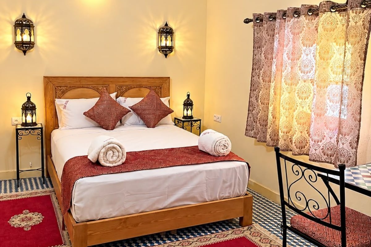 Quarto de hotel com cama de madeira, tapetes estampados, chão xadrez e arandelas decorativas.