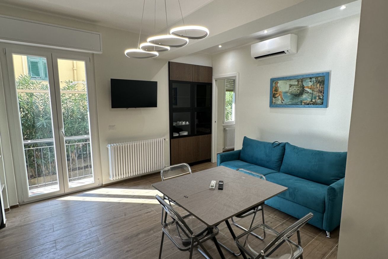 En moderne stue med en blå sofa, et spisebord med stole, et fladskærms-tv på væggen og et vindue med udsigt over grøn bevoksning.
