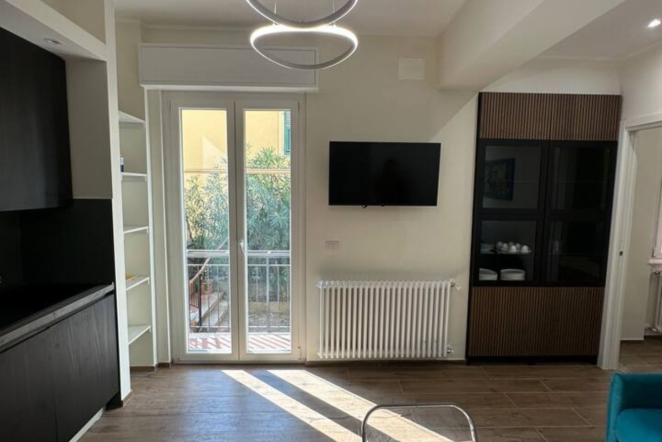 Una sala moderna con televisor de pantalla plana montado en la pared, puerta corrediza de vidrio que da a un balcón y un mueble empotrado con un televisor a la izquierda.