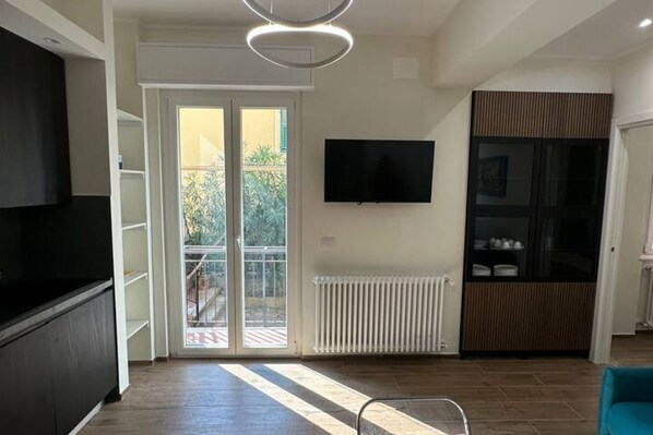 Een moderne woonkamer met een flatscreen televisie aan de muur, een schuifpui naar een balkon en een inbouwmeubel met daarin een televisie aan de linkerkant.