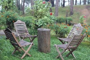 Dos sillas de madera rodean un tocón central, todo en un entorno de follaje verde y flores anaranjadas.