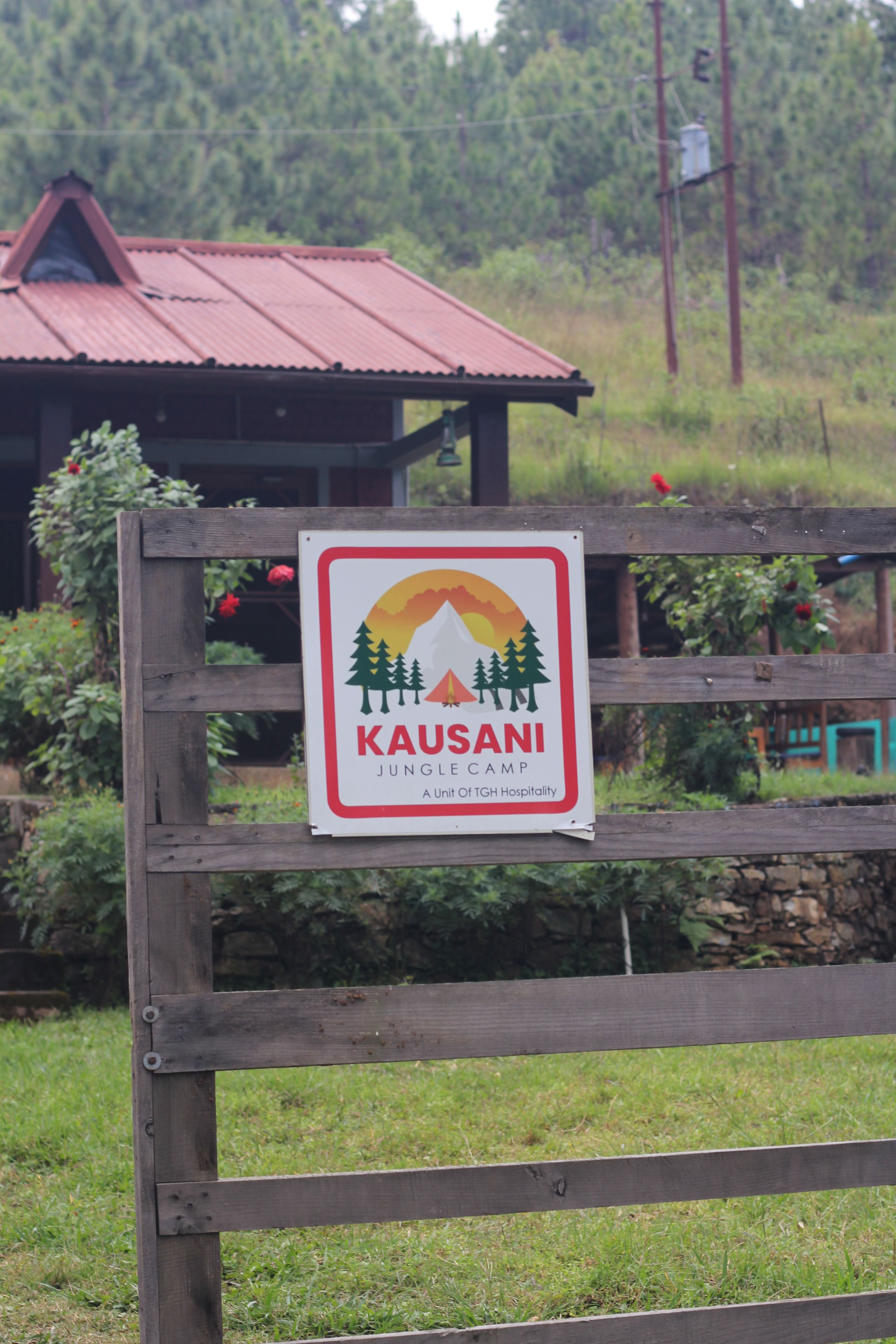 A sign for Kausani Jungle Camp, a unit of TGH Hospitality, with a wooden fence in the foreground.