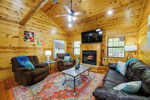 A cozy cabin interior with wooden walls, a fireplace, a ceiling fan, and a TV mounted on the wall. - Kickin Back Secluded Cabin - Hot Tub and Fire Pit (Broken Bow)