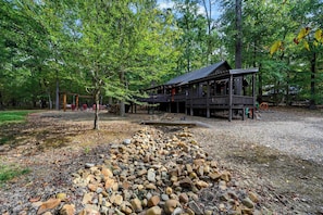 A rustic cabin with a porch, surrounded by a wooded area with a gravel path leading to it. - Kickin Back Secluded Cabin - Hot Tub and Fire Pit (Broken Bow)