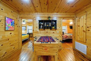 Um quarto aconchegante com paredes e pisos de madeira, mesa de pebolim, uma cama e uma prateleira com objetos decorativos.
