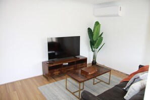Sala de estar con televisor de pantalla plana, mueble de entretenimiento de madera, mesa de centro, sofá y planta en maceta.