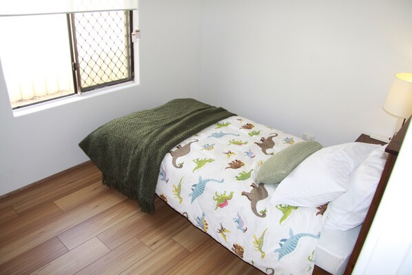 A small, single-bed room with a window, wooden flooring, and a bed with a dinosaur-themed bedspread.