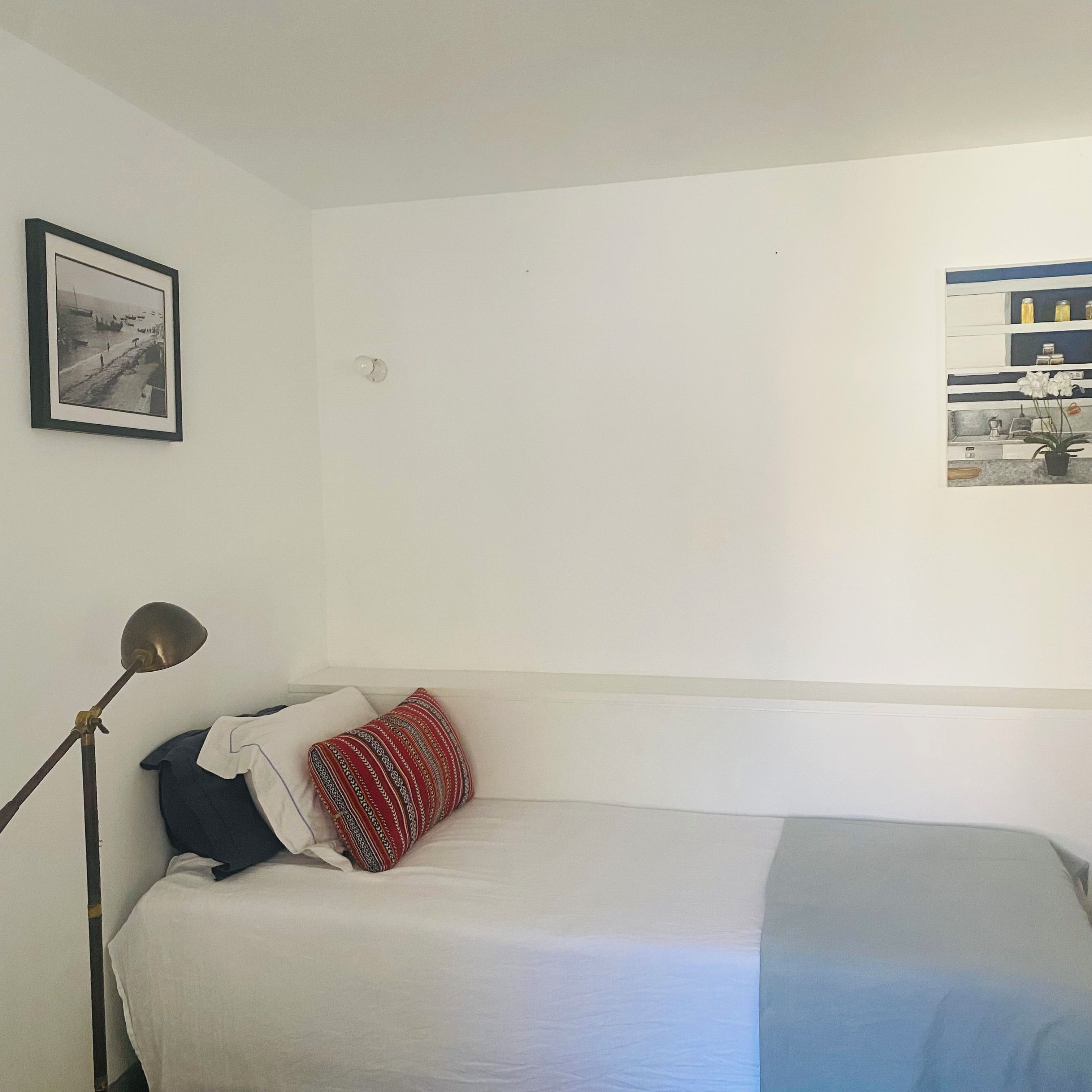Ein Einzelbett mit weißblauer Bettdecke, einem roten Kissen mit Muster und einer schwarzen Lampe, sowie ein gerahmtes Bild an der Wand.