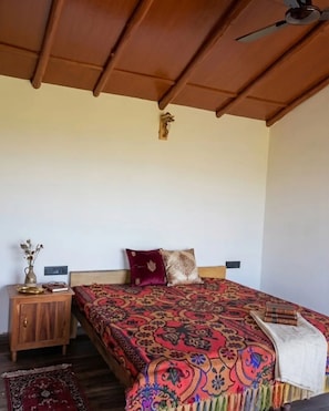 A bedroom with a wooden ceiling, a bed with patterned bedding, a nightstand with a vase and books, and a small rug on the floor. - Leela Orchard Sonapani (Mukteshwar)