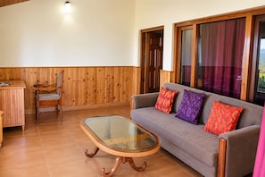 A room with a wooden wall, a glass-top table, a sofa with patterned cushions, and a chair. - Leela Orchard Sonapani (Mukteshwar)