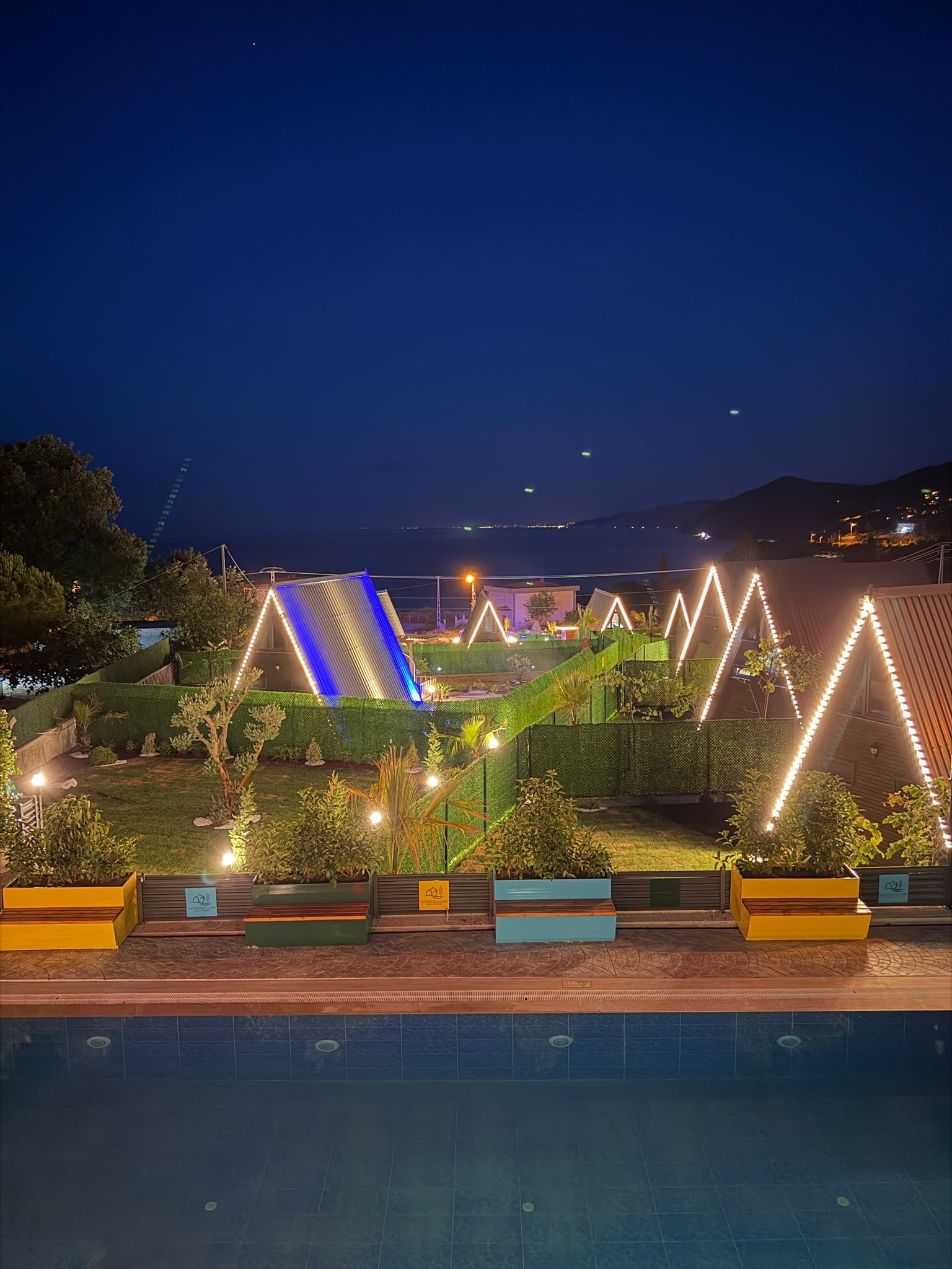 Vista nocturna d'una zona enjardinada amb piscina, edificis il·luminats i un paisatge urbà a la distància.