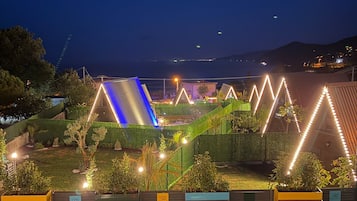 Vista nocturna d'una zona enjardinada amb piscina, edificis il·luminats i un paisatge urbà a la distància.