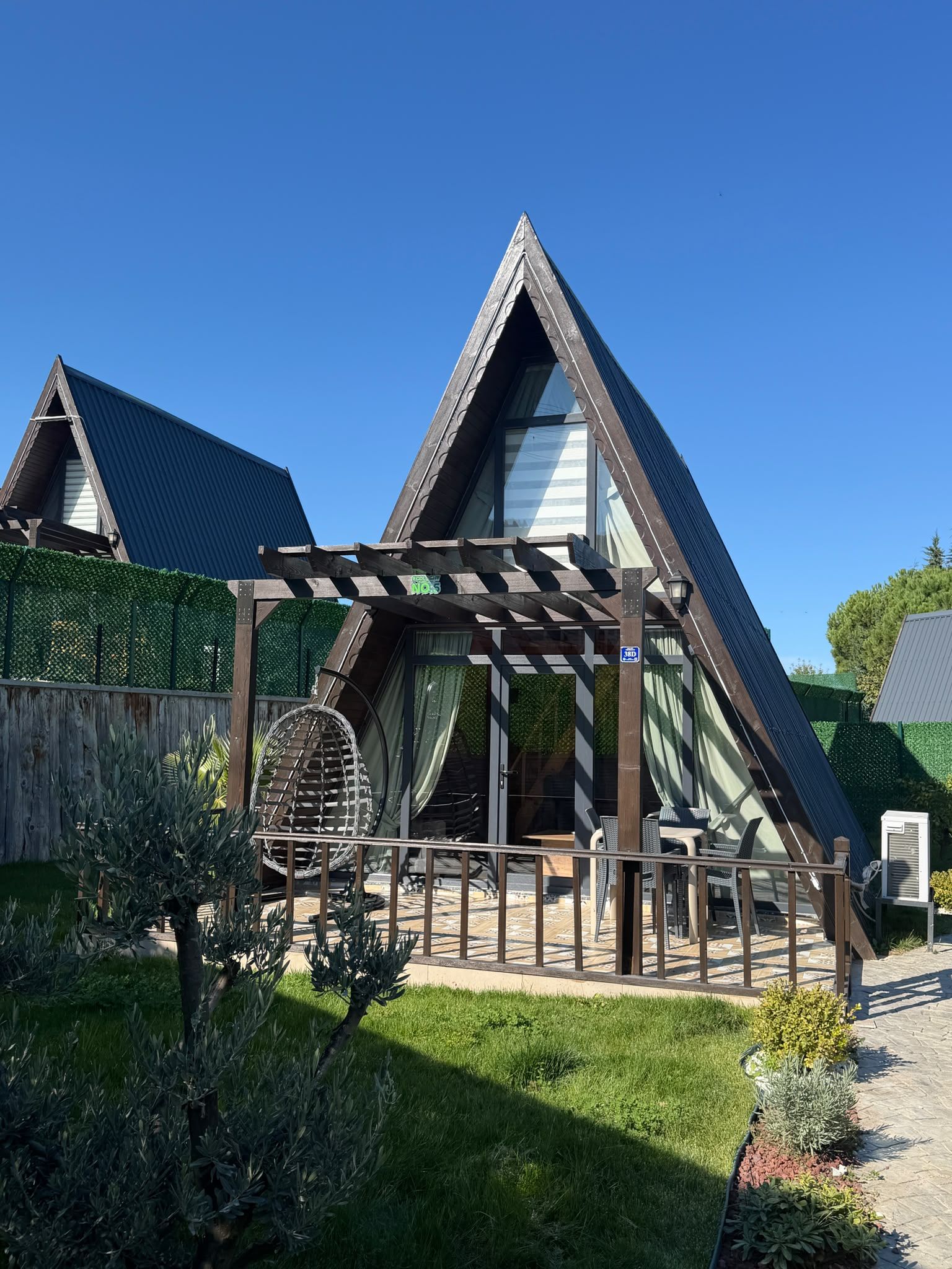 Una casa moderna con techo triangular, una pérgola de madera y un jardín con diversas plantas.