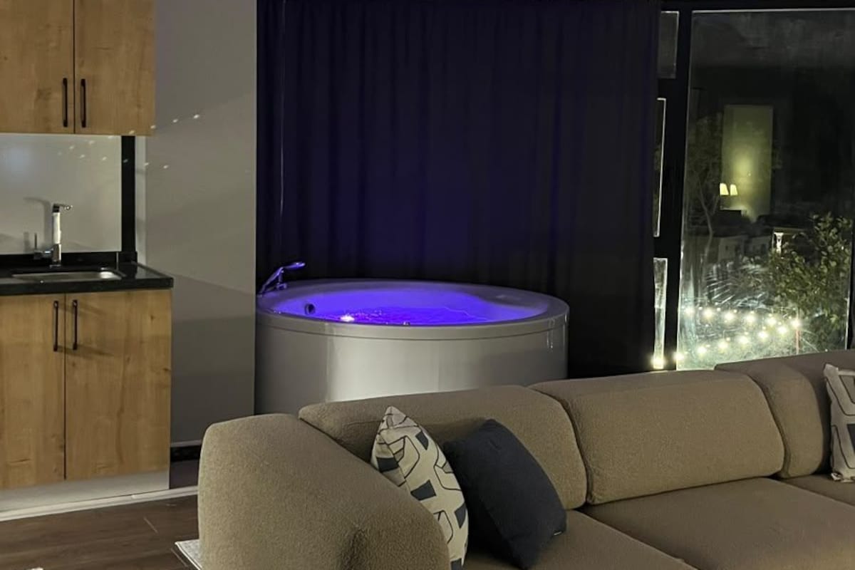 Ein modernes Wohnzimmer mit einer Couch, einem kleinen Tisch, einer Badewanne mit blauer Beleuchtung und Blick ins Freie.