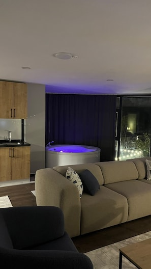 Una sala moderna con un sofá, una mesita, un jacuzzi con iluminación azul y vistas al exterior.
