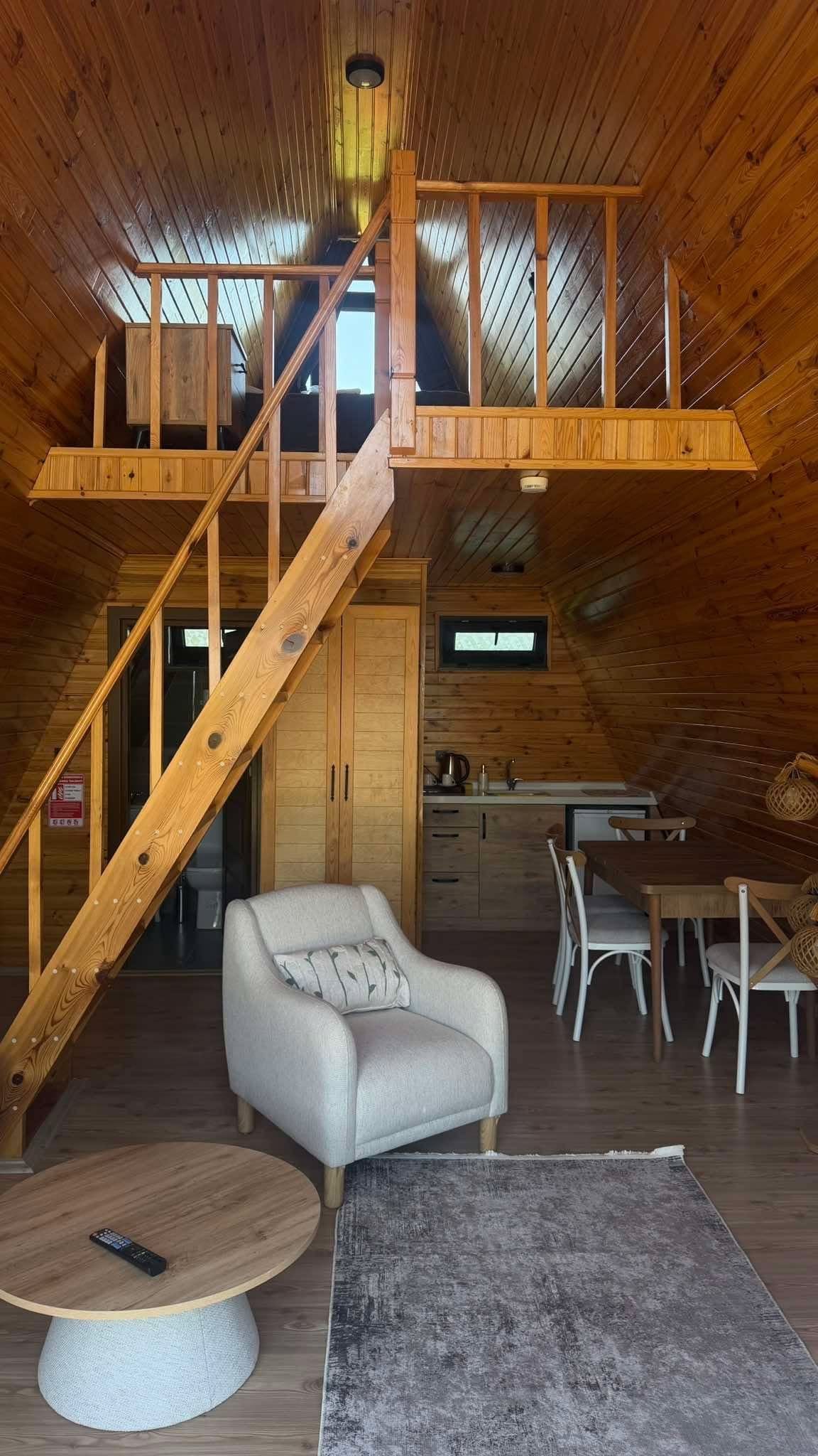 Een houten interieur met een trap naar een hoger gelegen verdieping, een bank, een eettafel met stoelen en een keukenhoek.