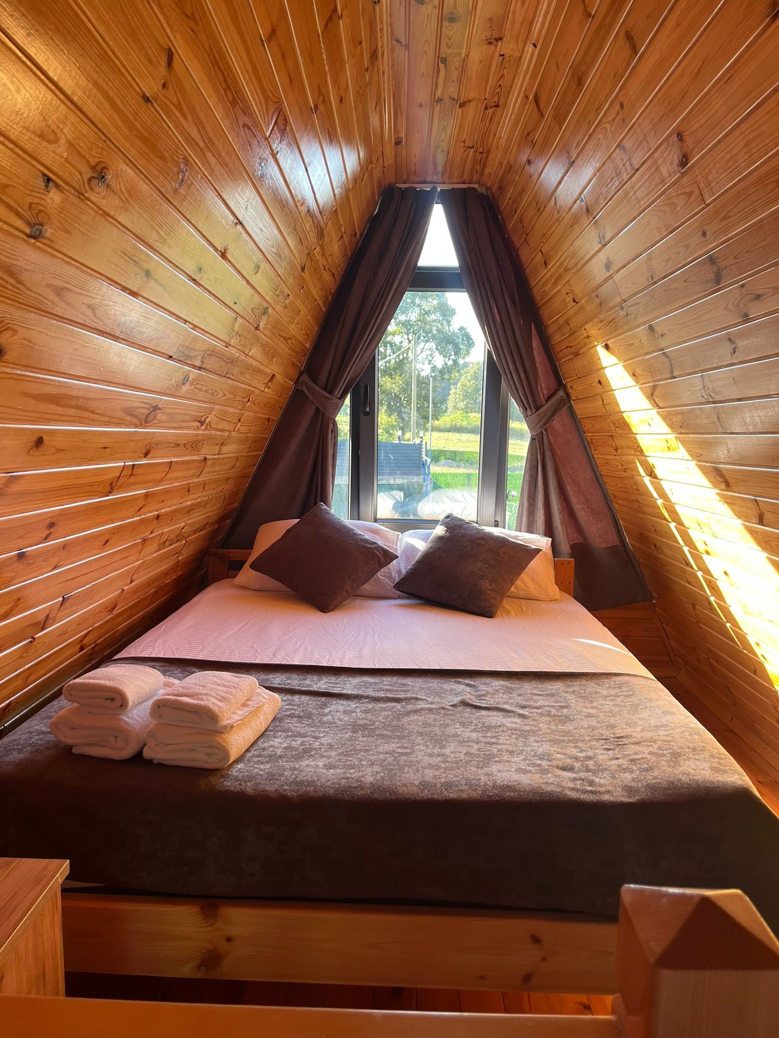 Una habitación pequeña y acogedora con paredes de madera y techo inclinado, que incluye una cama con almohadas y toallas dobladas, una ventana con cortinas y vistas al exterior con vegetación.