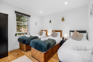 A hotel room with two beds, a window with a view of trees, and a small bedside table with a lamp. - Apartment Sleeps 7 With Wifi (London)