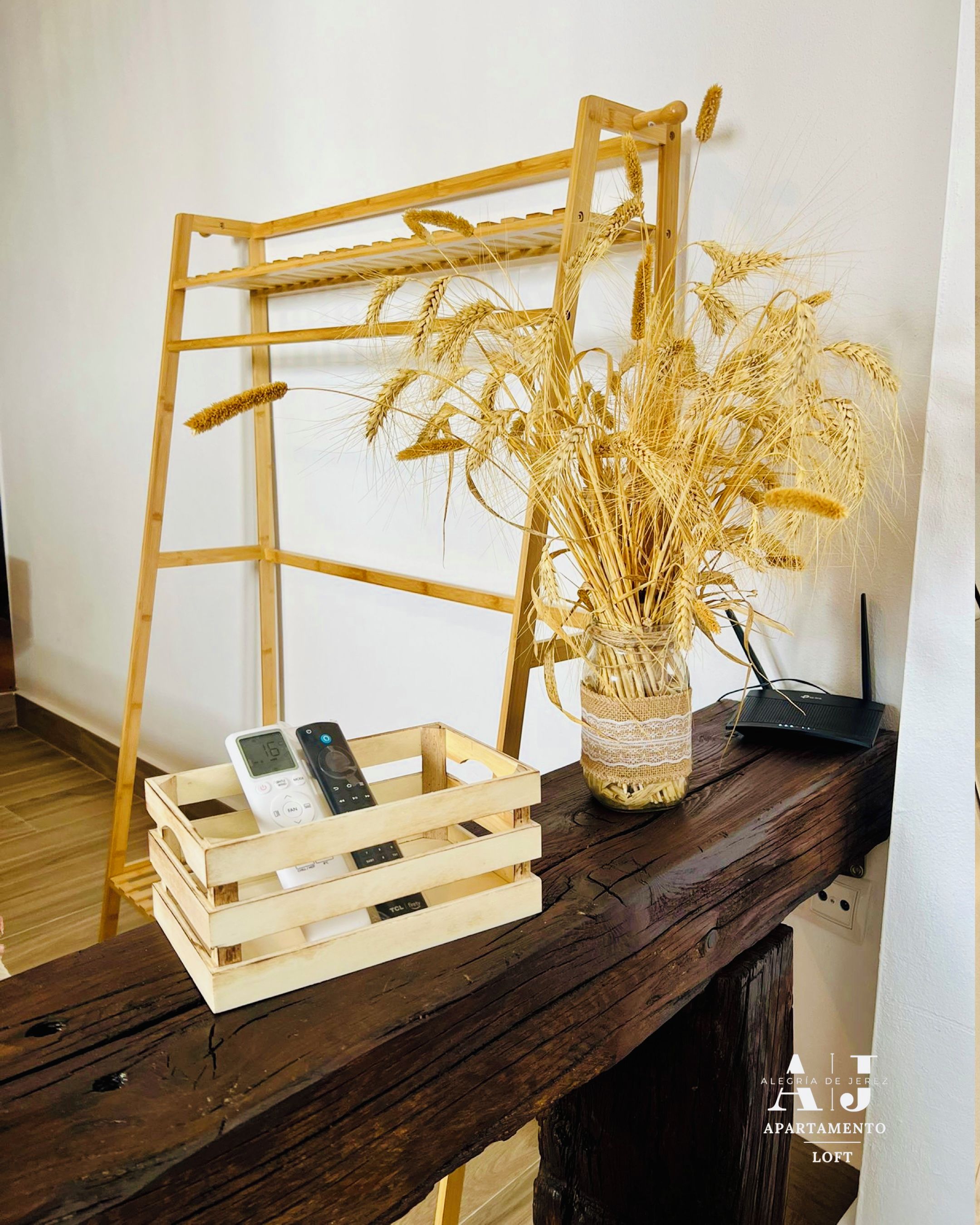Una escalera de madera apoyada en una pared, un jarrón con espigas de trigo secas y una caja de madera con un control remoto.