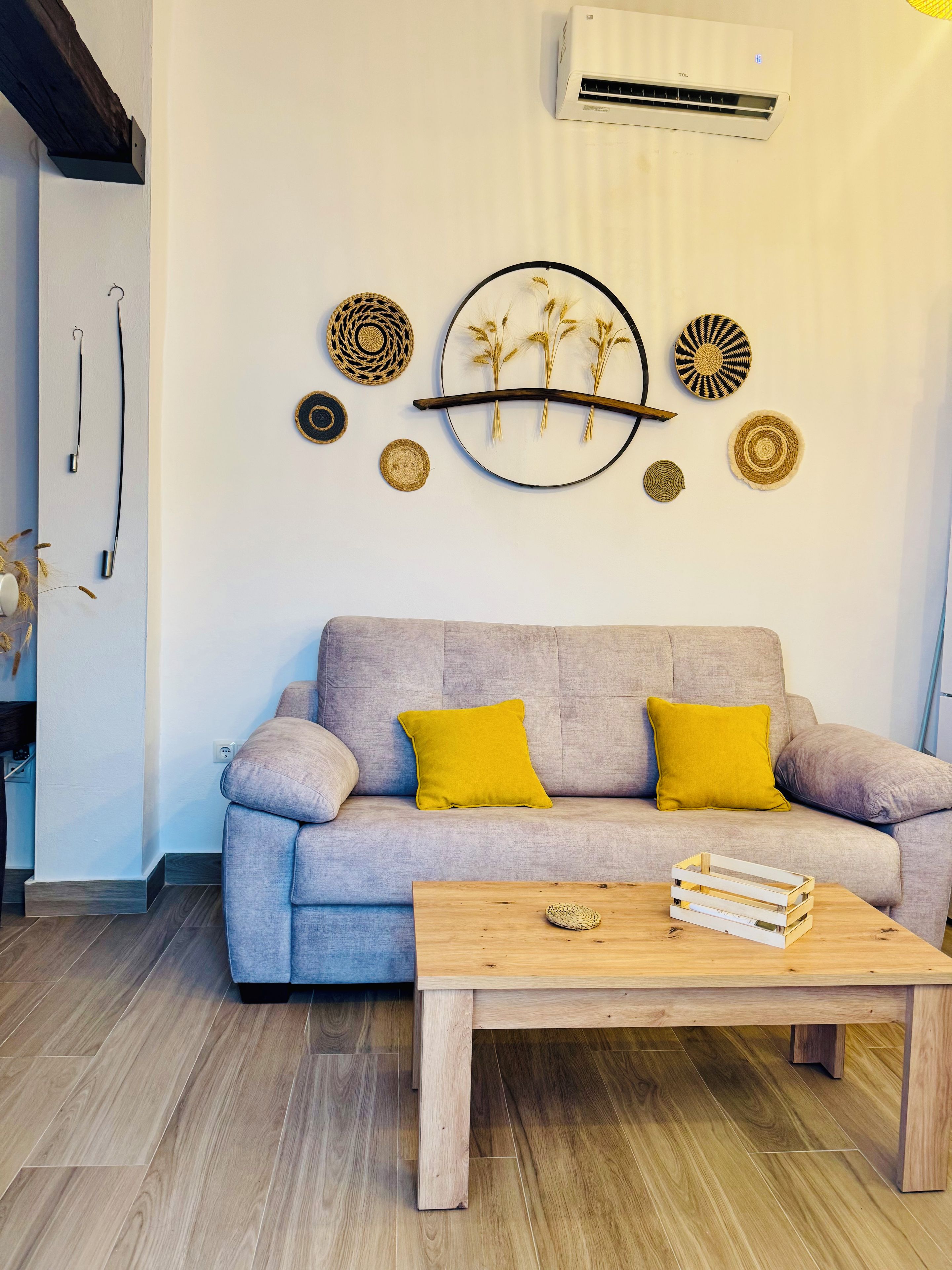 Una sala de estar con un sofá gris, dos cojines amarillos, una mesa de centro de madera y decoración en la pared que incluye platos tejidos y un plato circular con ramas.