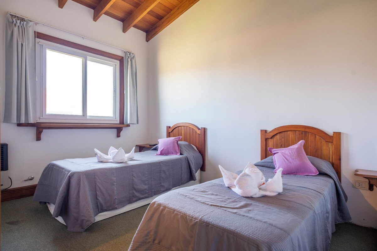 A room with two beds, each with a wooden headboard, a window with curtains, and a ceiling with exposed beams.