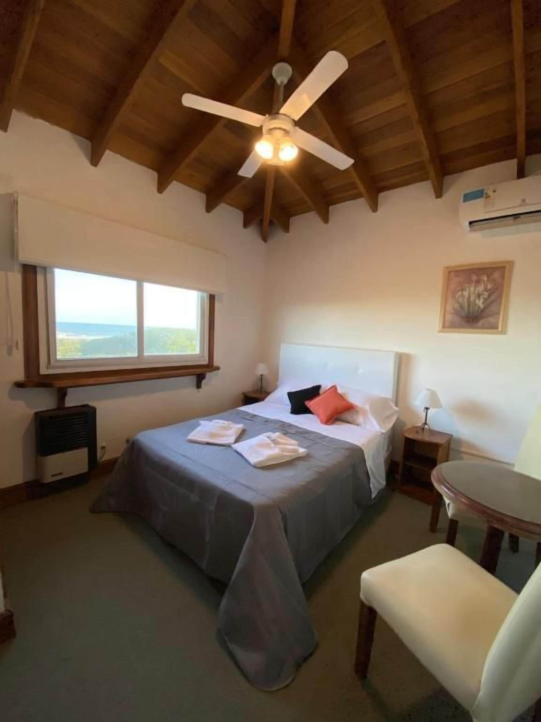 A bedroom with a bed, a chair, a desk, a ceiling fan, and a window.