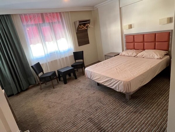 A hotel room with a bed, a desk, two chairs, a window with curtains, and a wall-mounted light fixture.