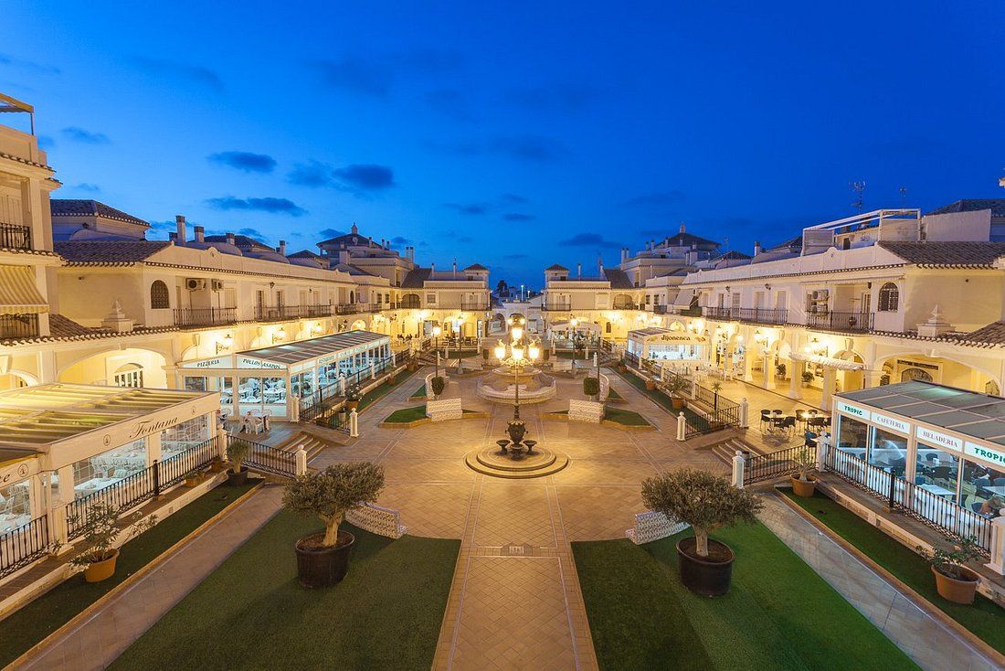 A courtyard with a central fountain, surrounded by buildings with outdoor seating areas and signage for restaurants.