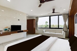 A modern hotel room with a large bed, a flat-screen TV, a wooden console, a white sofa, and a large window offering a view.