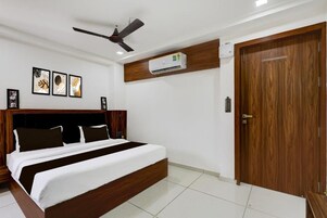 A bedroom with a bed, a ceiling fan, an air conditioning unit, a wooden door, and framed artwork on the wall.
