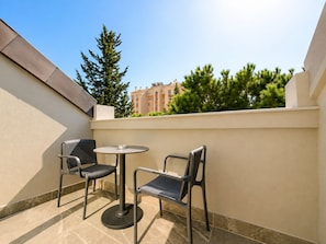 Zona d'estar exterior petita amb dues cadires i una taula, un paviment de rajola i vista a un edifici i arbres al fons.