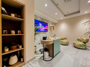 A modern reception area with a counter, a television displaying a colorful landscape, and a seating area with uniquely patterned chairs. - Leaf Lara Hotel (Antalya)